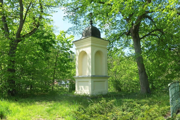 Kleines weißes Holocaust-Gedenkmonument in einem grünen Friedhof umgeben von einem Zaun und Bäumen unter einem klaren blauen Himmel in Vilnius, Litauen.