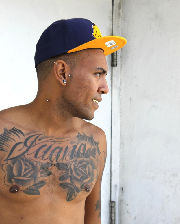 Eine Person mit einer Mütze mit einem Aufkleber mit einem Strichcode und Bildern, mit sichtbaren Tattoos auf dem Körper, die vor einer Tür und einer Wand steht.