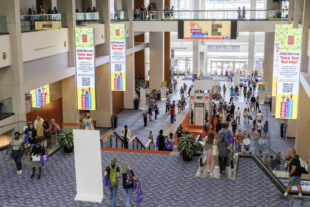 Eine große Gruppe von Menschen durchquert ein großes Gebäude mit Bannern, Topfpflanzen, Rolltreppen, Geländern, Säulen, Texttafeln und Glastüren, einige tragen Taschen, wahrscheinlich besuchen sie das National Book Festival.