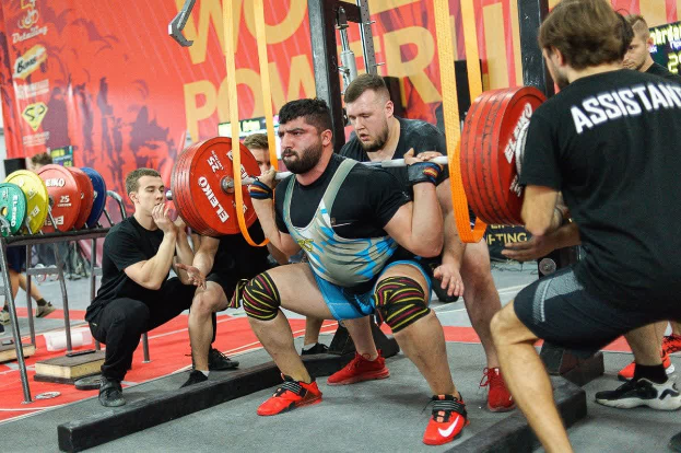 Eine Gruppe von Männern führt Kniebeugen mit Langhanteln in einem mit Geräten und Bannern vollgestellten Fitnessstudio durch.