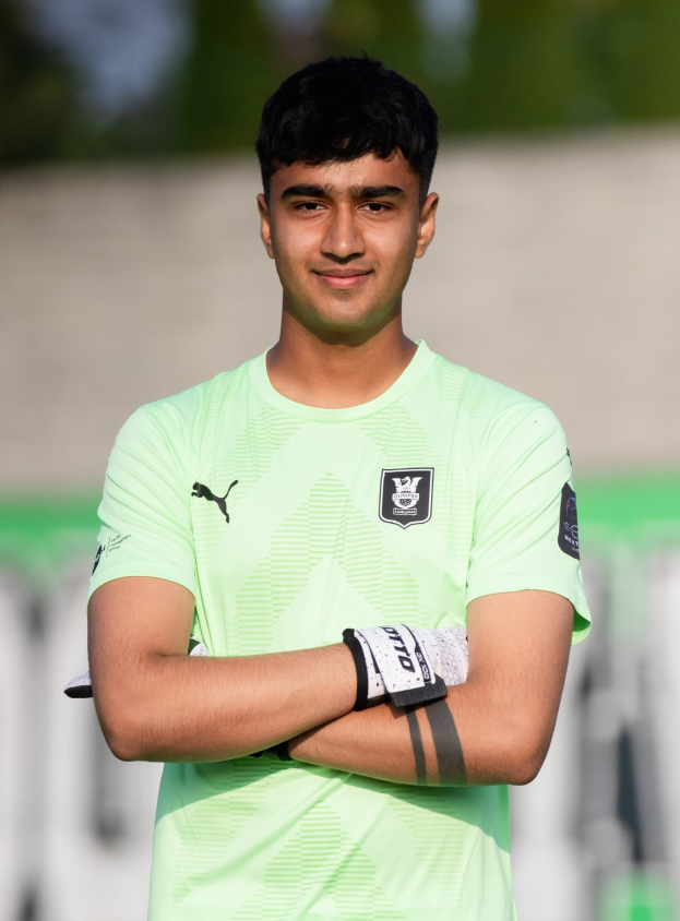 Ein junger Mann in einem grünen Hemd steht lächelnd auf einem Fußballfeld mit verschränkten Armen, der Hintergrund ist unscharf.