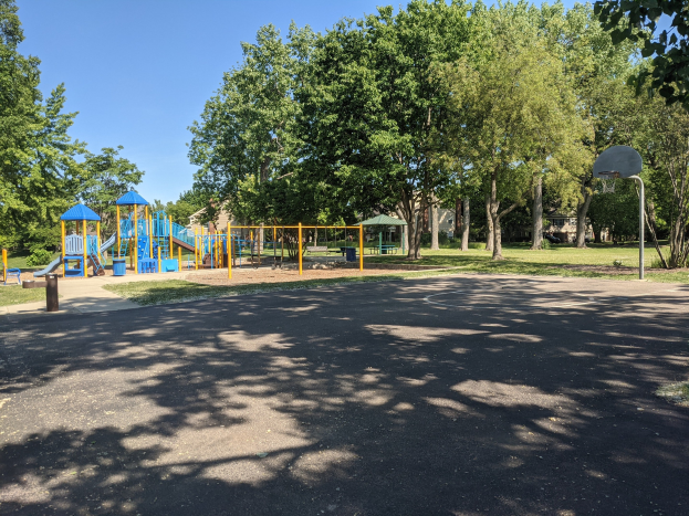 Ein Park mit einem Spielplatz in der Mitte, umgeben von Bäumen unter einem klaren blauen Himmel, eine Straße unten und ein Basketballkorb auf der rechten Seite.