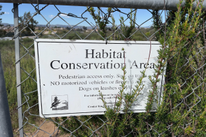 Ein Schild an einem Kettenzaun mit der Aufschrift "Habitat-Schutzgebiet" umgeben von Pflanzen, Bäumen, Hügeln und einem klaren blauen Himmel.