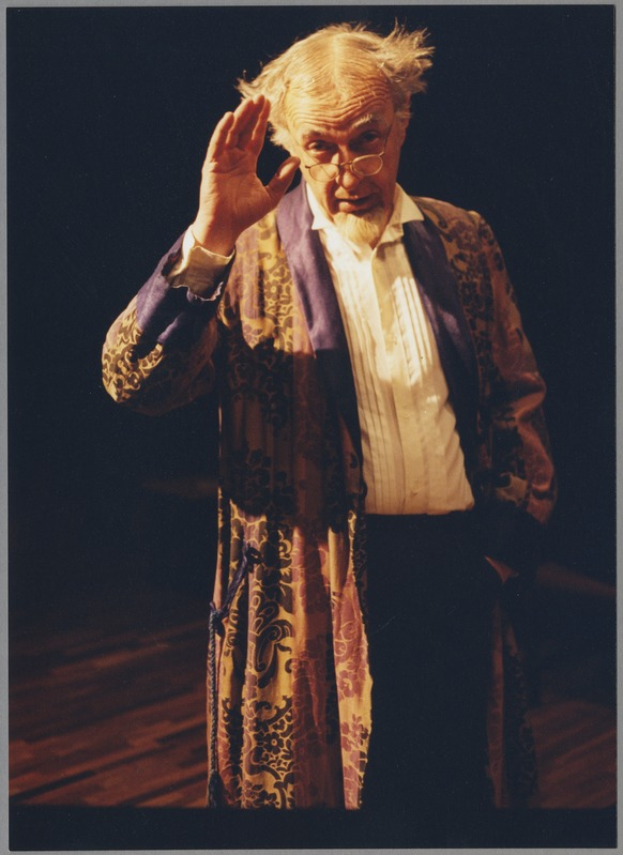 Ein Mann in einem Gewand und Brille steht auf einer Bühne mit dunklem Hintergrund, verschränkt die Hände und schaut ernst ins Publikum.