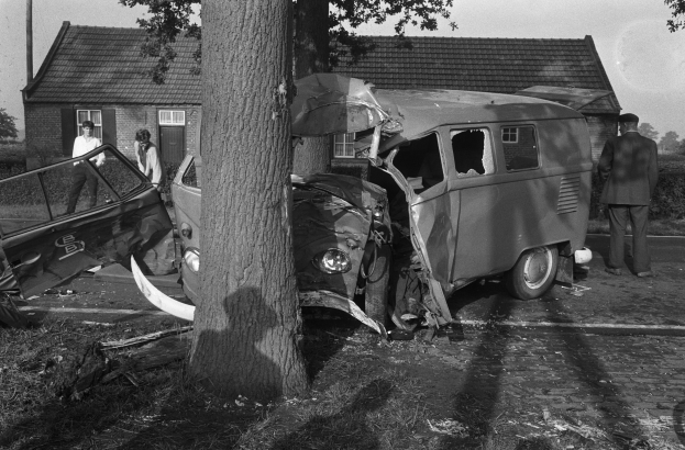 Eine Schwarz-Weiß-Szene eines verunglückten Autos am Straßenrand mit einem Baum im Vordergrund, einigen Menschen in der Nähe und Häusern, Bäumen und einem Pfahl im Hintergrund.