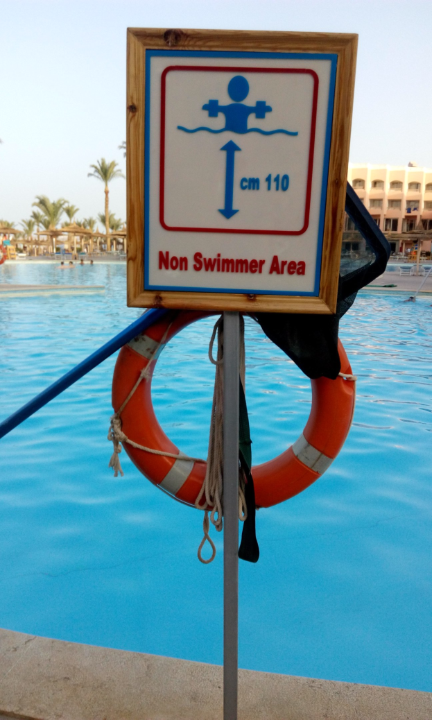 Ein Schwimmbad mit einem "Nichtschwimmerbereich"-Schild und einem Rettungsring an einem Pfahl, umgeben von Bäumen, Gebäuden und einem klaren blauen Himmel.