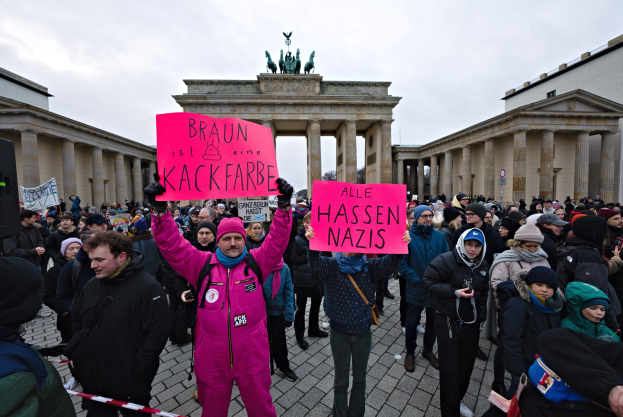Eine Gruppe von Menschen demonstriert vor dem Reichstag in Berlin, hält Schilder gegen die Anti-Nazis-Bewegung der deutschen Regierung und zeigt Gebäude, Säulen und Statuen im Hintergrund bei klarem blauem Himmel.