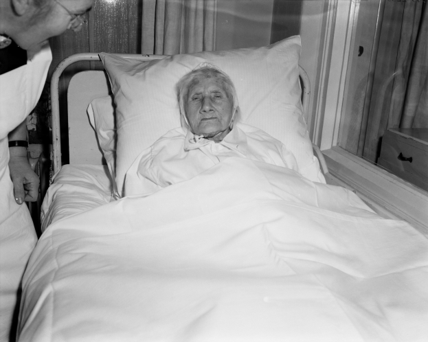 Eine ältere Frau liegt in einem Krankenhausbett mit einer weißen Bettdecke und einem Kissen, während eine Krankenschwester neben ihr steht, mit einem Fenster und Vorhängen im Hintergrund.