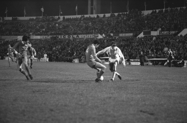 Schwarzes und weißes Bild von Männern, die Fußball spielen, in einem Stadion mit Zuschauern auf den Rängen und Fahnen im Hintergrund.