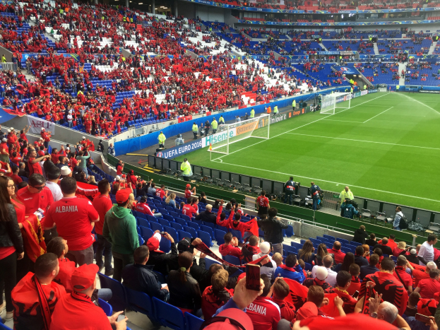 Eine große Menschenmenge in einem Stadion schaut einem Fußballspiel zu, mit Menschen auf Sitzen und Stehen, Bannern, einem Torpfosten und Deckenbeleuchtung.