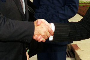 Zwei Männer im Business-Outfit schütteln sich die Hand vor einem Gebäude, umgeben von grünem Gras und Pflanzen, was eine formelle Geschäftsbesprechung suggeriert.