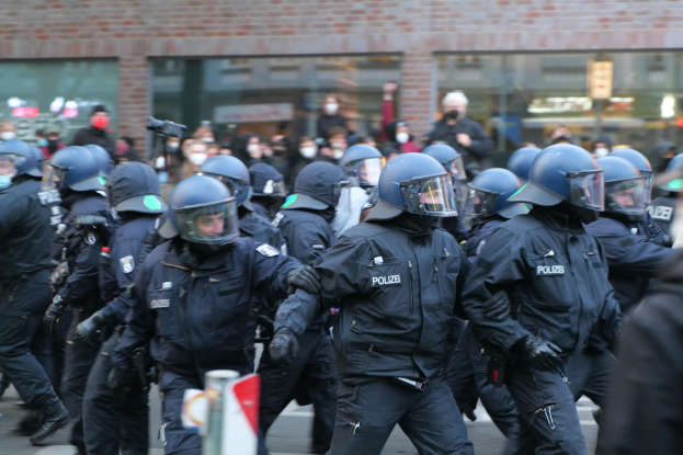 Eine Gruppe von Polizisten in Einsatzausrüstung, einschließlich Helmen und Handschuhen, die eine Straße entlanggehen mit einem unscharfen Hintergrund, der ein Gebäude mit Glaswänden und ein Schild an einem Pfosten zeigt.