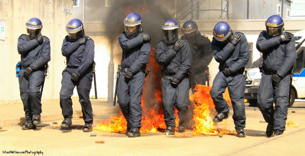 Menschen in Helmen stehen vor einem Feuer mit Gegenständen auf dem Boden, Gebäuden im Hintergrund, einem Fahrzeug, einem Plakat und einer Tafel an der linken Wand und Text am unteren Bildrand.