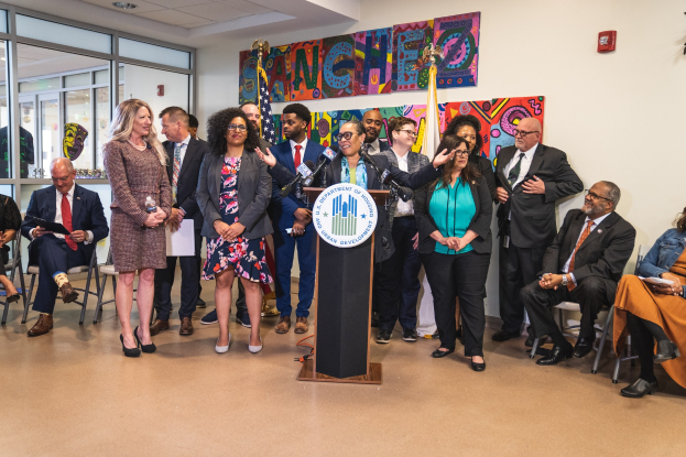 Eine Gruppe von Menschen steht in der Nähe eines Podiums mit einem Mikrofon und einer Tafel mit der Aufschrift "Sancho City Councilman Joins City Council", während andere auf Stühlen sitzen oder im Raum stehen, mit Flaggen, einem Feuerlöscher und Deckenleuchten.