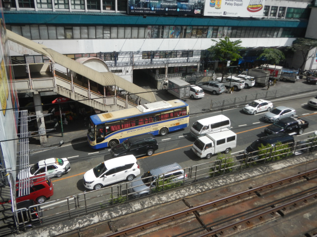 Eine belebte Stadtstraße mit Fahrzeugen, Gebäuden, Grünflächen und verschiedenen städtischen Elementen einschließlich Schienen und Beschilderung.