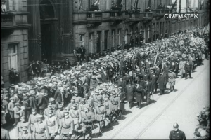 Schwarzes Foto einer Parade mit einer großen Menge, die eine Straße entlang marschiert, einige halten Gewehre in den Händen, vor einem Gebäude mit einem Wasserzeichen in der rechten oberen Ecke.
