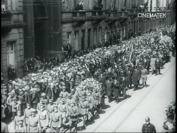 Schwarzes Foto einer Parade mit einer großen Menge, die eine Straße entlang marschiert, einige halten Gewehre in den Händen, vor einem Gebäude mit einem Wasserzeichen in der rechten oberen Ecke.
