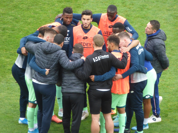 Eine Gruppe von M%C3%A4nnern in Jacken, die sich auf einem Fussballfeld im Kreis versammelt haben.