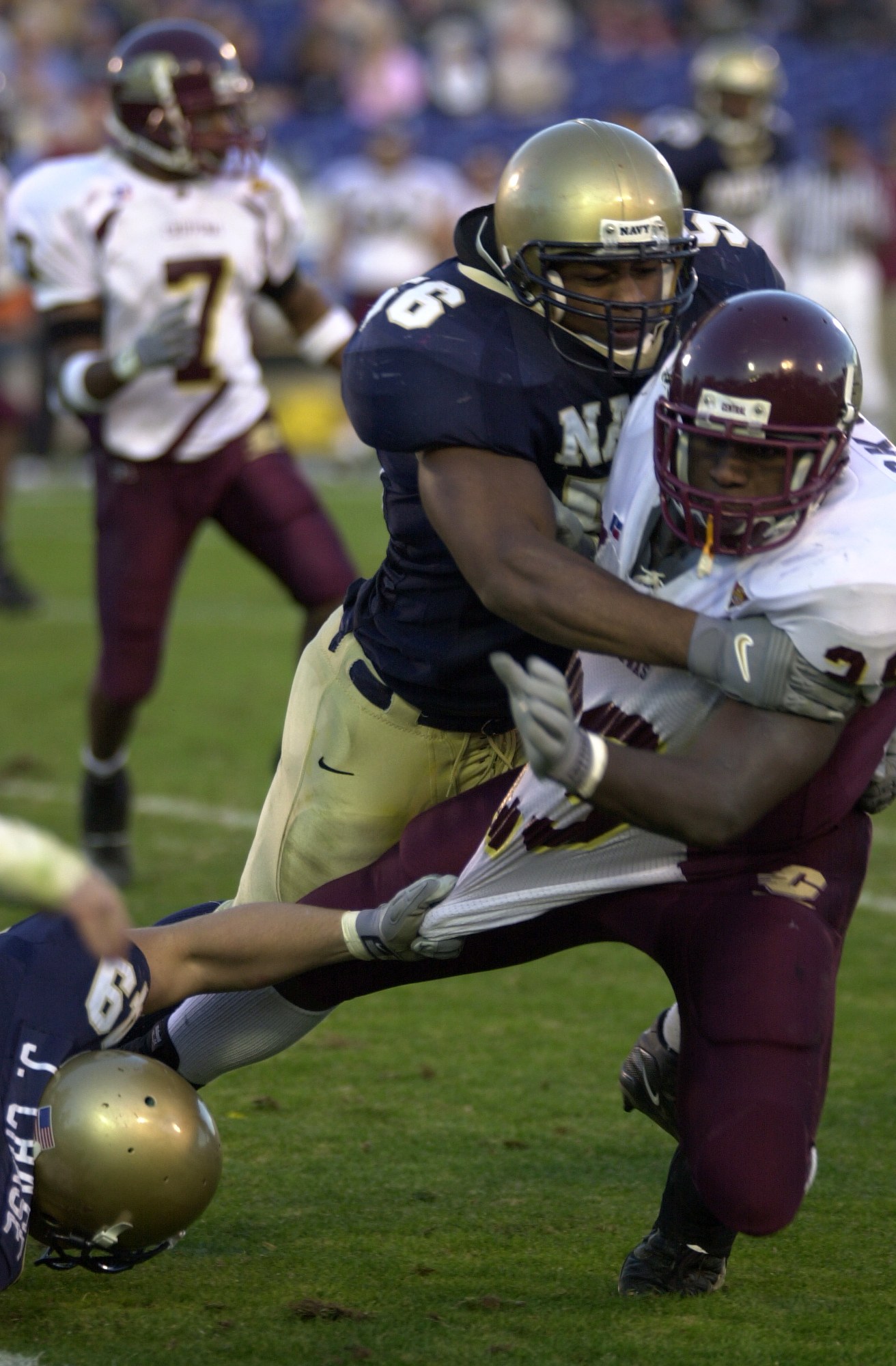 Zwei Fußballer in Sportkleidung und Helmen stoßen auf dem Feld zusammen, während andere zuschauen.