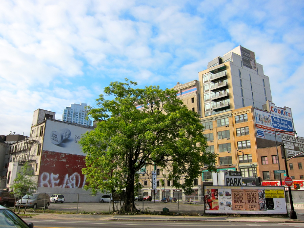 Stadtansicht mit mehreren Gebäuden, einer Straßenlaterne, einem Baum und mehreren geparkten Autos unter einem leicht bewölkten blauen Himmel.