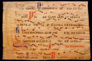 Ein illuminiertes Manuskript mit Musiknotation auf einem Blatt Papier vor einem dunklen Hintergrund, gefüllt mit Text und Musiksymbolen, die ein komplexes Design bilden.