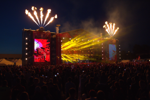 Eine große Menschenmenge auf einem Musikfestival mit einer Bühne, auf der Künstler auftreten und von hellen Lichtern beleuchtet wird, umgeben von Bannern, Zelten links und einem feuerwerksbeleuchteten dunklen Himmel im Hintergrund.