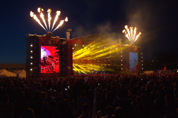 Eine große Menschenmenge auf einem Musikfestival mit einer Bühne, auf der Künstler auftreten und von hellen Lichtern beleuchtet wird, umgeben von Bannern, Zelten links und einem feuerwerksbeleuchteten dunklen Himmel im Hintergrund.