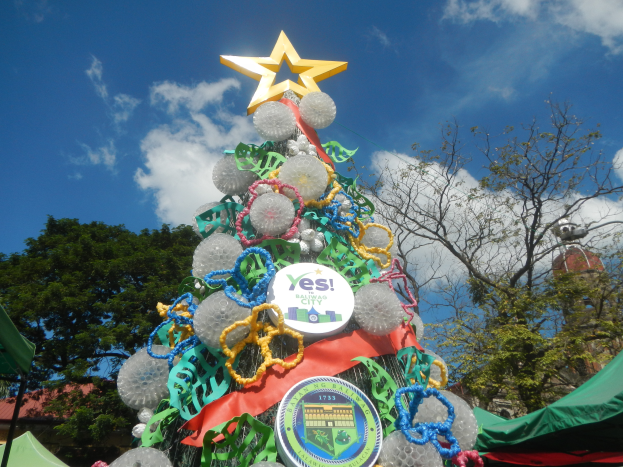 Ein Weihnachtsbaum mit einem Stern darauf, umgeben von bunten Schleifen und Dekorationen, mit Zelten, Bäumen, Gebäuden und einem klaren blauen Himmel im Hintergrund.
