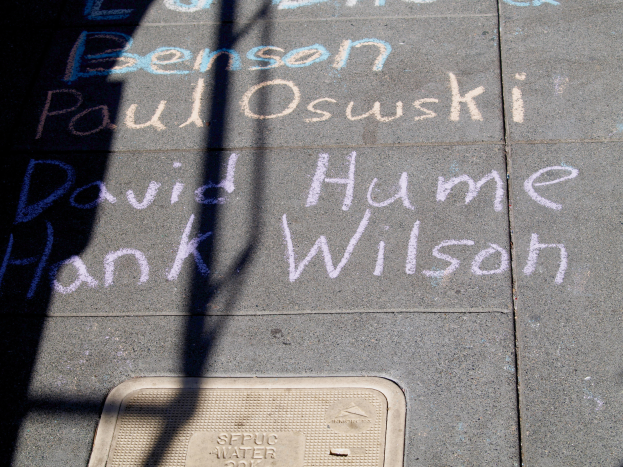 Ein Gehweg mit Kreide-Schrift, die wie ein Denkmal aussieht, ein Gullydeckel unten und ein Schatten eines Baums auf der linken Seite.