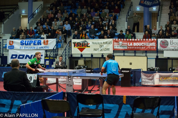 Zwei Männer spielen Tischtennis drinnen, mit mehreren Zuschauern, die stehen und sitzen, einigen leeren Stühlen und Werbetafeln, mit Text in der linken unteren Ecke.