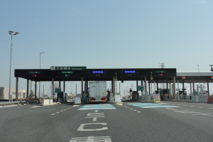 Eine Mautstation mit einem vor ihr geparkten Lastwagen, Straße mit weißen und blauen Markierungen, Laternenpfähle, Schilder, Gebäude, Bäume und einen klaren blauen Himmel im Hintergrund.