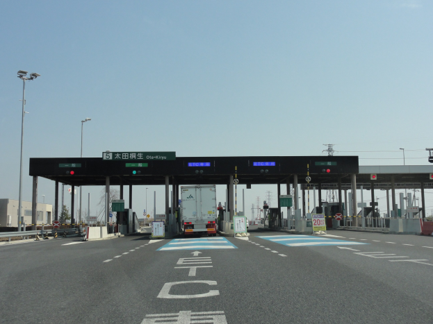Eine Mautstation mit einem vor ihr geparkten Lastwagen, Straße mit weißen und blauen Markierungen, Laternenpfähle, Schilder, Gebäude, Bäume und einen klaren blauen Himmel im Hintergrund.