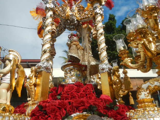 Ein Rosen- und Goldschmuck verzierter Umzugswagen mit Rosenstatuen und -blüten, im Hintergrund ein Gebäude, Bäume und ein bewölkter Himmel.