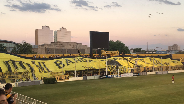 Ein Fußballfeld mit einer Gruppe von Menschen darauf, umgeben von einem Metallzaun, einem Torpfosten, einer Fahne mit der Aufschrift "Boca Juniors vs Real Sociedad", einigen Pflanzen, einem Anzeigebildschirm, einigen Pfählen, der Rinde eines Baumes, einigen Bäumen, einigen Gebäuden und einem bewölkten Himmel.