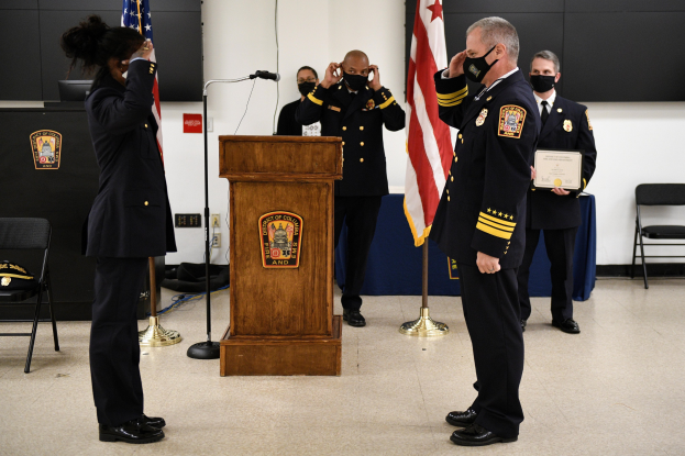 Gruppe uniformierter Personen in Masken auf einem Podium während einer Vereidigung eines neuen Feuerwehrchefs, der eine Urkunde hält, mit Flaggen, Stühlen, Tischen und einer Wand im Hintergrund.