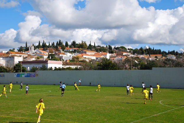 Gruppe von Menschen, die draußen ein Spiel spielen, mit Plakaten an einer Wand, Bäumen, Strommasten, Häusern und Wolken im Hintergrund.