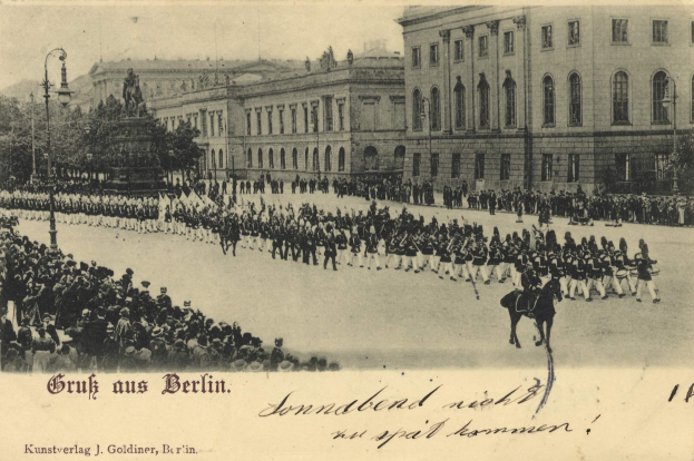 Ein altes Schwarz-Weiß-Foto eines Umzugs in Berlin, Deutschland, mit Menschen auf dem Boden und auf Pferden, einer Statue auf einem Sockel, Straßenlaternen, Bäumen, Gebäuden mit Fenstern und einem bewölkten Himmel, mit Text am unteren Bildrand.