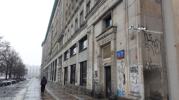 Eine Person geht an einem Gehweg vorbei an einem Gebäude mit Graffiti, mit parkenden Autos auf der Straße und Bäumen auf beiden Seiten, während Gebäude und Himmel im Hintergrund zu sehen sind.