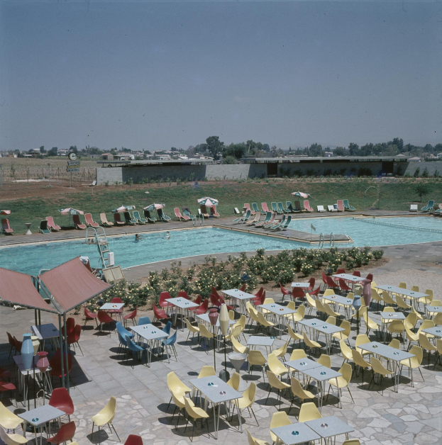 Ein Freibad mit Tischen, Stühlen und Sonnenschirmen umgeben von Blumenpflanzen, einem Zaun, einer Wand, Bäumen, Gebäuden und einem bewölkten Himmel.