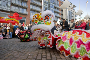 Ein lebendiges chinesisches Neujahrsfest in Amsterdam mit einer Löwen-Tanzdarbietung im Vordergrund, umgeben von einer Menschenmenge und Gebäuden im Hintergrund unter einem klaren blauen Himmel.
