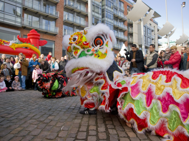 Ein lebendiges chinesisches Neujahrsfest in Amsterdam mit einer Löwen-Tanzdarbietung im Vordergrund, umgeben von einer Menschenmenge und Gebäuden im Hintergrund unter einem klaren blauen Himmel.