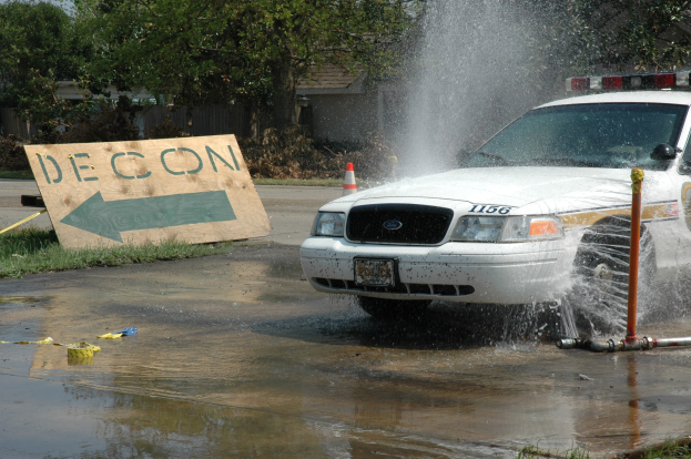 Ein Streifenwagen fährt durch eine Pfütze, mit einem Schild, einer Verkehrskegel und anderen Gegenständen am Boden, mit Bäumen, Gebäuden und einem klaren blauen Himmel im Hintergrund.