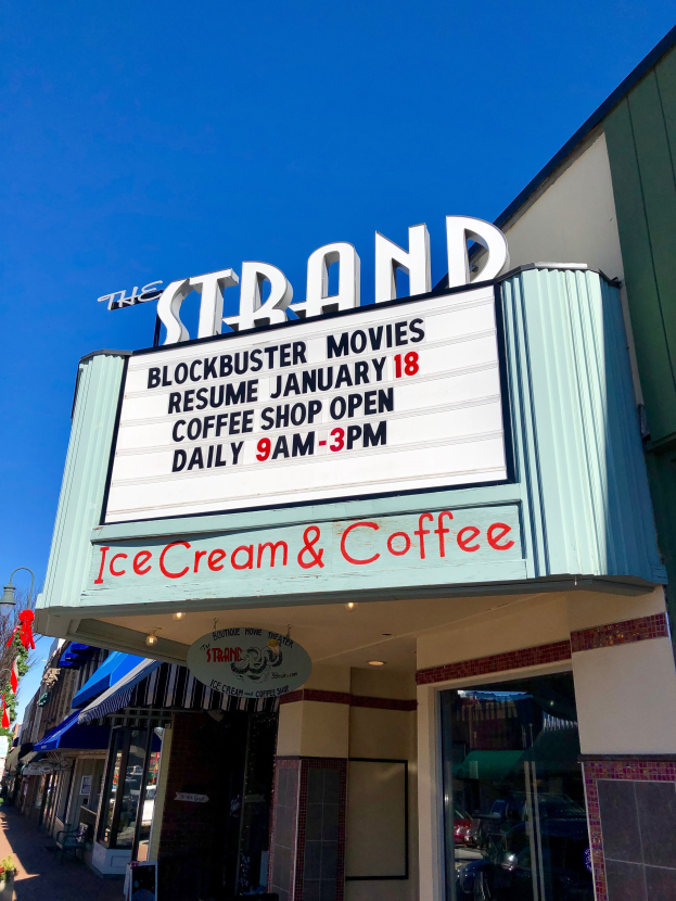 Der Strand Blockbuster Movies Resume Januar 18 Kaffeehaus mit einem Namensschild, einem Straßenschild, einer Baumgruppe, Topfpflanzen und einer bewölkten Himmel.
