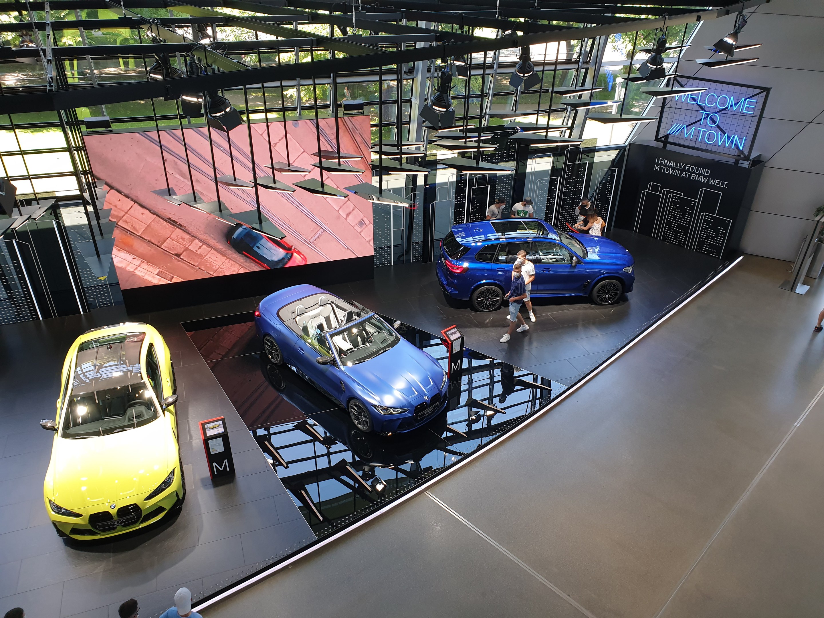 Eine Gruppe von Menschen steht auf dem Boden des BMW Museums in München, Deutschland, mit Autos zur Linken und einem Schild mit der Aufschrift "BMW Museum München" zur Rechten, mit Blick auf Bäume, Gebäude und den Himmel durch Glasfenster.