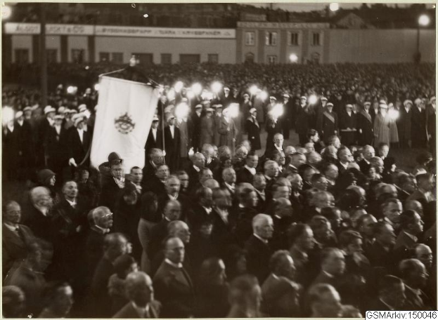 Schwarzes und weißes Foto einer großen Menge vor einem Gebäude, mit einer Person, die eine Fahne hält, in der Mitte, Stangen und Lichter im Hintergrund und Text unten mit der Aufschrift "Gustav Klimts Rede an der Berliner Mauer".