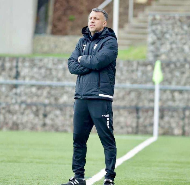 Ein Fussballtrainer auf einem Feld mit Überkreuzten Armen, der eine schwarze Jacke und Hose trägt, vor einem Zaun, Treppe und Wand.