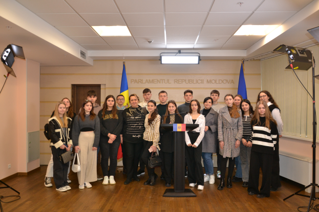 Eine Gruppe von Menschen steht vor einem Podium mit einem Mikrofon, umgeben von Fahnen, einer Wand mit dem Text "Moldova Jugendkongress 2018", einer Tür, einem Dach mit Deckenleuchten und Ständern mit Lichtern.