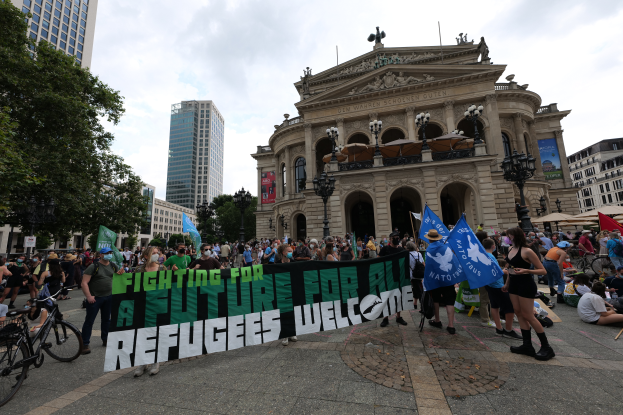 Eine Gruppe von Menschen, die vor einem Gebäude steht und sitzt, hält ein Banner mit der Aufschrift "Kämpfen für eine Zukunft für Flüchtlinge Willkommen", einige halten Fahnen, Fahrräder, Laternenpfähle, Bäume und Gebäude sind im Hintergrund zu sehen, der Himmel ist bewölkt.