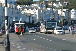 Eine belebte Stadtstraße mit hohen Gebäuden, Fahrzeugen, Fußgängern, Ampeln, Laternen, Texttafeln, Bäumen und einem klaren blauen Himmel.