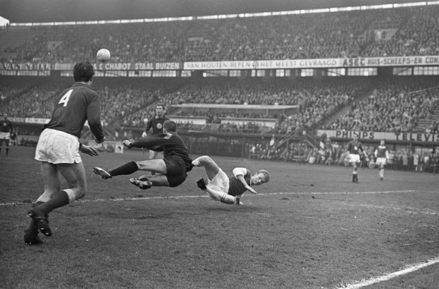 Eine Gruppe von Männern, die Fußball auf einem Feld in einem Stadion spielen, mit Zuschauern auf den Tribünen und Transparenten mit Text im Hintergrund, dargestellt in Schwarz-Weiß.